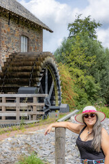 Wall Mural - Happy smiling female tourist standing in front of old Eper or Wingbergermolen water mill, hat, sunglasses, tree on blurred background, sunny day in Terpoorten, Epen, South Limburg, Netherlands