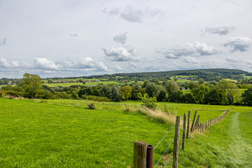 Wall Mural - Dutch agricultural panoramic landscape, wooden posts and wire fences dividing plots, trees, hills, villages against cloud covered sky in background, sunny day in Epen, South Limburg, Netherlands