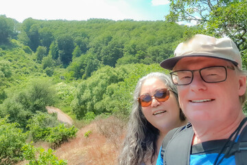 Wall Mural - Smiling senior adult couple taking a selfie on mountain with abundant leafy trees in background, hiking trail in Wallfhärte Weidingen nature reserve, sunny summer day in Utscheid, Germany