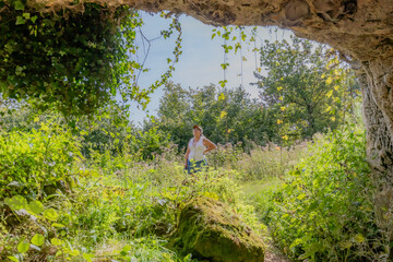 Wall Mural - View from inside a cave senior female hiker standing surrounded by wild leafy vegetation, Thier de Lanaye nature reserve in Belgian part of Sint-Pietersberg, sunny day with blue sky in Vise, Belgium