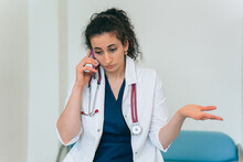 Puzzled brunette female doctor in medical gown with phonendoscope talks by phone with misunderstanding face expression. Tired female doctor exhausted. Medicine, pandemic.