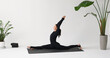 A yoga instructor performs the Hanumanasana exercise, longitudinal splits with arms raised up, training in a black tracksuit while sitting on a mat in the studio