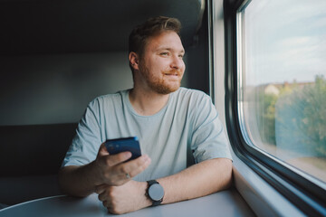 A man with a beard and mustache in a blue t-shirt is using a smartphone while traveling by Railway train, sitting in the train and looking out the window.