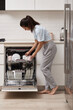 happy woman unloading plate from open automatic built-in dishwasher machine with clean utensils inside in modern white kitchen.