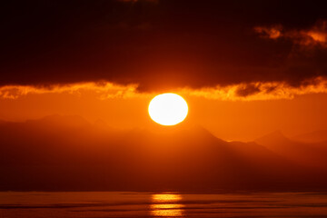 Telephoto image of large setting sun overtop a fjord mountain in Norway.  Beautiful orange sun rising up from the peaks of Norwegian mountains.  Shot from the top of Reinebringen