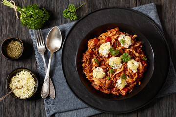 Wall Mural - Lasagnette with beef ragu, mushrooms and ricotta cheese in black bowl on dark wooden table with herbs and seasoning, italian recipe, horizontal view from above