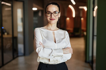 Waist up portrait of smiling Caucasian brunette businesswoman in glasses crossing arms and looking at camera standing in office building