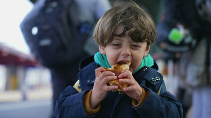 Portrait of a delighted happy child eating croissant standing at train platform wearing jacket and scarf during autumn fall season. Kid snacking carb food with wide smile