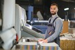 © Serhii - Printing house interior with modern offset print machine and operator in working uniform checking quality and controlling process of print