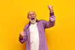 old bald grandfather in purple shirt celebrates victory with his mouth open on yellow isolated background
