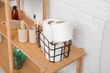 Basket with toilet paper rolls and bath supplies on wooden shelving unit near light brick wall, closeup