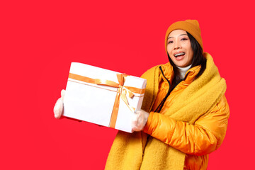 Poster - Young Asian woman in winter clothes with gift on red background