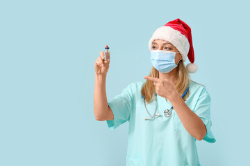 Poster - Mature female doctor in Santa hat and medical mask with ampule on blue background