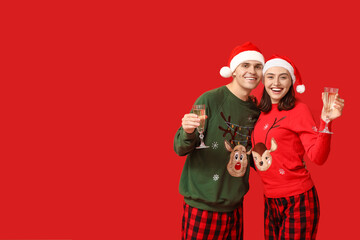 Wall Mural - Happy young couple in Christmas pajamas and Santa hats drinking champagne on red background