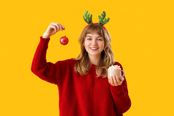 Canvas Print - Pretty young woman holding cup of hot chocolate and Christmas ball on yellow background