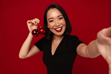 Wall Mural - Pretty asian woman smiling while taking selfie with Christmas tree toys isolated over red background