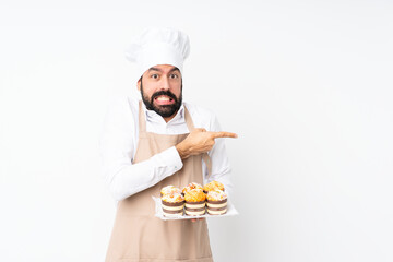 Wall Mural - Young man holding muffin cake over isolated white background frightened and pointing to the side