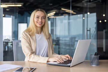 Portrait of a young businesswoman sitting in the office at the desk, working on a laptop, smiling and looking at the camera