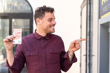 Poster - Young caucasian man taking a lot of money at outdoors pointing to the side to present a product