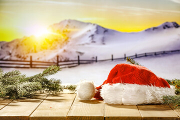 Canvas Print - Wooden table in light color.Christmas tree branches on the top of the table for decoration.Red Santa Claus hat.Free space for your products and gifts Blurry landscape of mountains covered with snow.