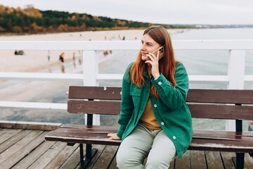 Wall Mural - Young happy 30s woman tourist talking on smart phone and sitting on the bench outdoors. Redhead Woman walking along the waterfront, seaside. High quality photo