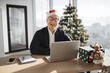 © sofiko14 - Asian businessman wearing glasses sitting in office near xmas tree looking at camera and working on laptop during the holiday season, typing on computer keyboard.