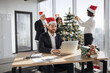 © sofiko14 - Attractive Caucasian businessman employee in suit and santa hat sitting at desk using laptop in christmas decorated office. Young corporate worker in festive workplace looking at screen of computer.