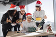 © sofiko14 - Secret Santa. Smiling Asian businessman in Santa hat unpacks a Christmas present from multinationals colleagues at work standing around him. New Year's eve.