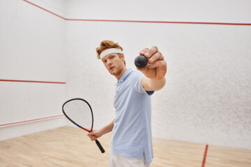 Wall Mural - redhead man in sportswear showing squash ball and holding racquet while standing inside of court