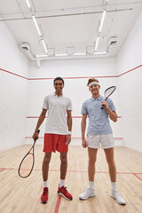 Wall Mural - vertical shot, happy interracial male friends looking at camera and holding racquets for squash