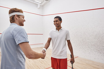 Wall Mural - joyful interracial sportsmen in active wear smiling and shaking hands while holding squash racquets