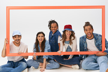 Wall Mural - happy and stylish interracial friends sitting and posing with picture frame on grey backdrop