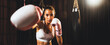 Asian female Muay Thai boxer punch fist in front of camera in ready to fight stance posing at gym with boxing equipment in background. Focused determination eyes and prepare for challenge. Spur