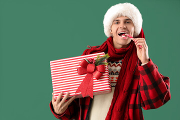 Sticker - Happy young man in Santa hat with Christmas gift and candy cane on green background