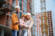 Man tossing up apartment keys and smiling while standing next to wife and daughter outside building under construction. Happy family homeowners posing on the street at construction site.