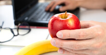 The Concept Of A Healthy Snack At Work. A Young Man Is Holding An Apple And Working On A Laptop At Home Or At Work. Close-up. Selective Focus.
