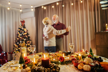 selective focus on christmas table with ornaments and food with blurry senior couple dancing on chri