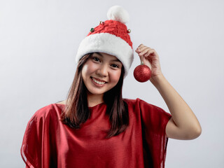 A portrait of a joyful Asian woman celebrating Christmas, dressed in a red outfit and Santa hat, holding Christmas accessories like ornaments and playing with them. Isolated against a white background