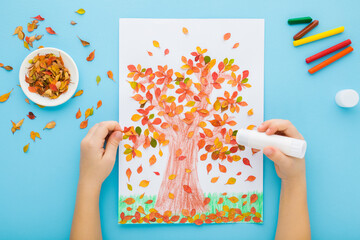 Little child hands gluing and creating tree shape from colorful leaves on white paper. Light blue table background. Pastel color. Making autumn decorations. Point of view shot. Closeup. Top down view.