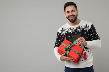Wall Mural - Young man in Christmas sweater opening gift on grey background. Space for text
