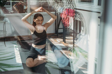 Woman With Arms Raised Doing Yoga On Mat At Home Seen Through Glass