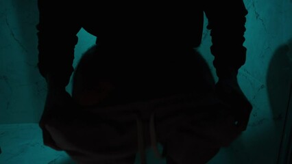 A man enters a dark bathroom with dim blue lighting, takes off his shorts, and sits on the toilet.
