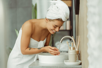 Portrait of young cheerful woman wearing white towel after shower, washing face with water