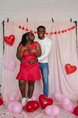 Wall Mural - Full length of happy young African couple embracing on pink background with heart shaped balloons