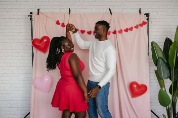 Wall Mural - Romantic young African couple dancing on pink fabric background with heart shaped balloons