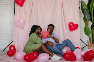 Wall Mural - Smiling African couple covering faces with heart shaped Valentine card while sitting on 