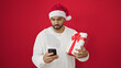 © Krakenimages.com - African american man holding christmas gift using smartphone over isolated red background