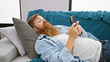 Poster - Handsome bearded irish redhead man, fully relaxed and serious, focused on using his phone, lost in a world of internet tech as he lies on the sofa at home, texting away in his cozy living room.