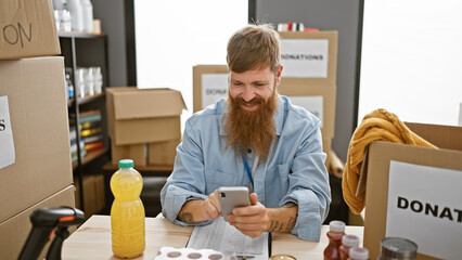 Canvas Print - Confident young redhead man volunteering with joy at charity center, smiling while typing on smartphone sitting at table