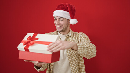 Wall Mural - Young hispanic man wearing christmas hat unpacking gift over isolated red background